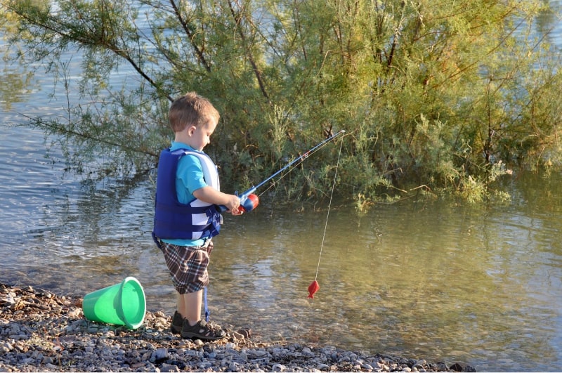 a boy with a fishing pole and a green pail near a river