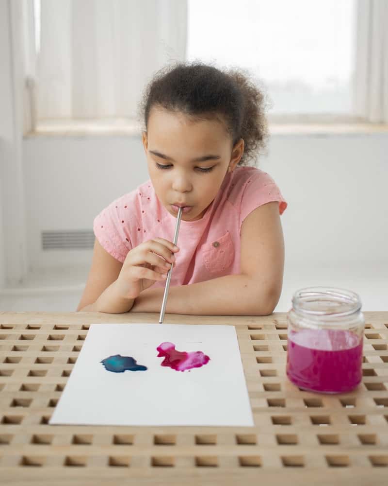 a girl painting with a straw and watercolor paints
